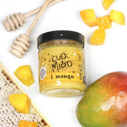 Słoik miodu z serii cud z mango 250g firmy Sądecki Bartnik. Słoik leży w otoczeniu plastra miodu, mango, nabieraków. Etykieta na słoiku ma napis: "Cud miód i mango" oraz informacjami o produkcie. Zakrętka jest w kolorze czarnym.