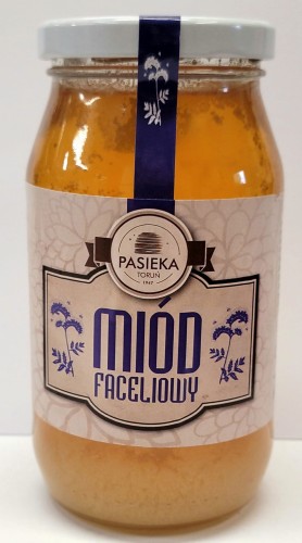 Miód Faceliowy 0,6k g - Pasieka Toruń. Przezroczysty słoik z żółtą zawartością, jasna etykieta z fioletowymi literami oraz kwiatami facelii umieszczona w brązowej ramce  zawierająca logo, nazwę, producenta, pojemność oraz skład. Biała zakrętka.