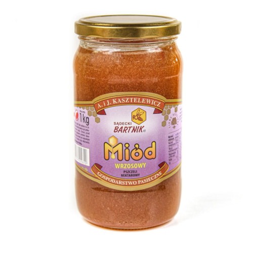 Miód Wrzosowy 1 kg Sądecki Bartnik. Etykieta w fioletowym kolorze zawierająca nazwę produktu, wagę, producenta . Miód o jasno brązowej barwie w przezroczystym słoiki ze złota nakrętka.