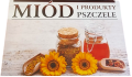 Tablica informacyjna z plexi z obrazkiem produktów pszczelich: miodu, pyłku oraz propolisu i pomarańczowych kwiatków. Powyżej jest brązowymi literami napis: miód i produkty pszczele" przedzielone linią i brązową wstawką. Tło jest szare.