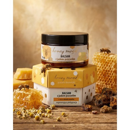 Balsam z jadem pszczelim Honey Mood o pojemności 100ml, który stoi na podniesieniu, czyli pudełku w które jest zapakowany słoiczek z balsamem. Pudełko ma heksagonalny kształt. W otoczeniu są pszczoły, propolis, miód oraz suszone kwiaty.