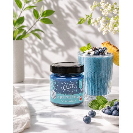 Miód ze spiruliną serii "Cud" o pojemności 250g. Miód w słoiku jest w kolorze niebieskim z czarną zakrętką. Obok miodu jest koktajl przygotowany z proponowanego miodu. W tle są kwiaty oraz rozsypane borówki.